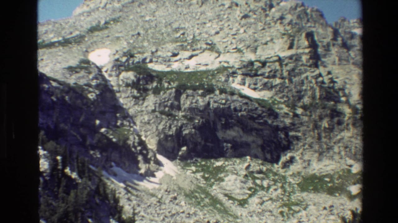1980 Grand Teton home-movie frame showing a steep rocky slope with snow remnants, green alpine growth, and conifers.