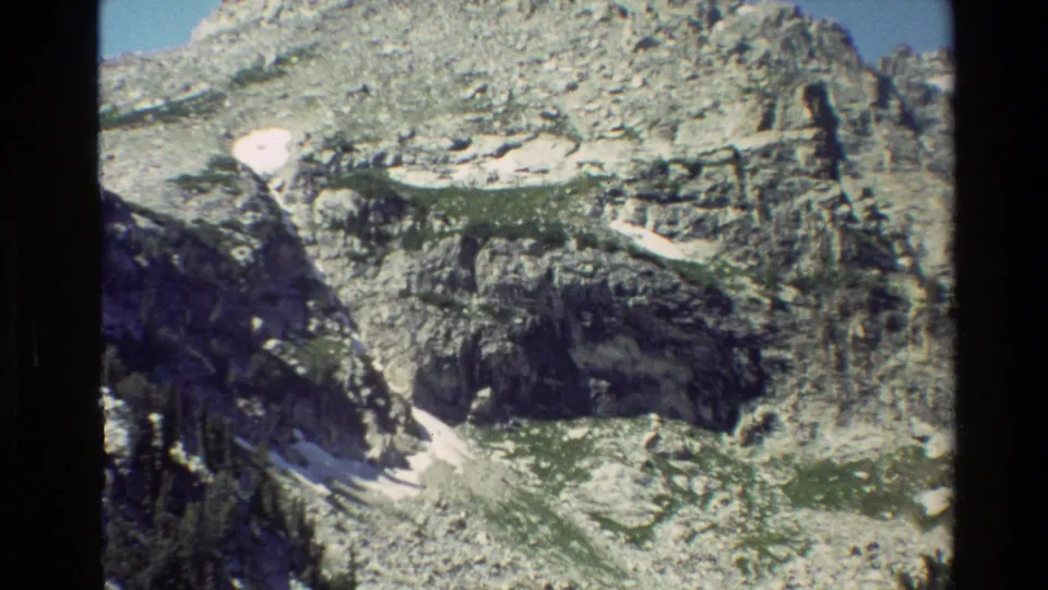 1980 Grand Teton home-movie frame showing a steep rocky slope with snow remnants, green alpine growth, and conifers.