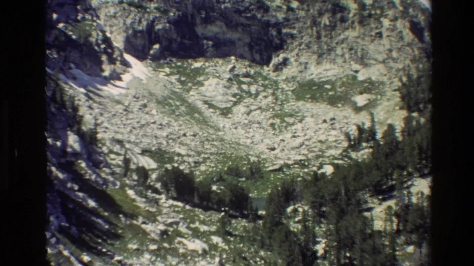 1980 Grand Teton footage frame showing rugged rock, alpine vegetation, conifers, and a small shaded snow patch.