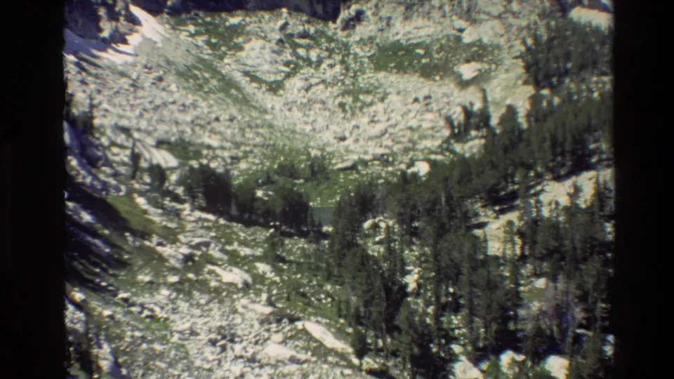 Grand Teton 8mm frame from 1980 showing a lower alpine slope with talus, conifers, mossy vegetation, and small snow patches.