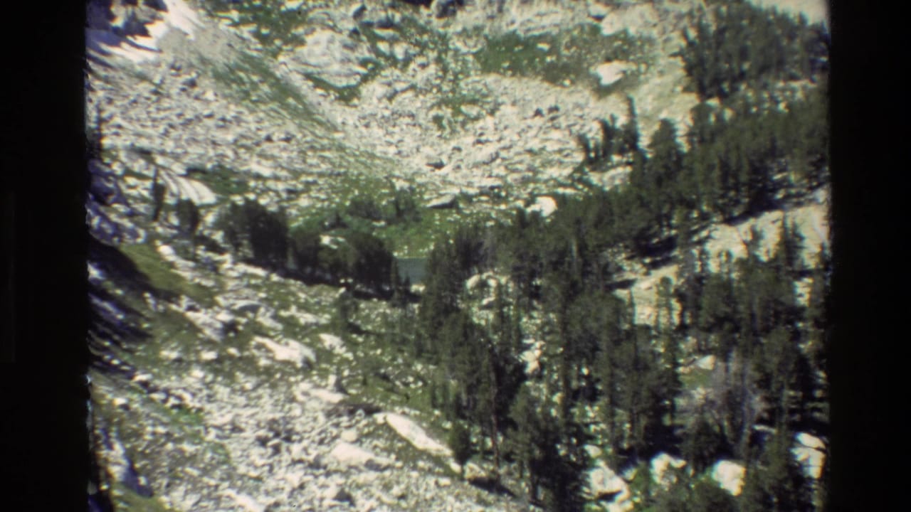 1980 8mm aerial view in Grand Teton, Wyoming, showing rocky mountain terrain, conifer stands, and green-blue lake water below.