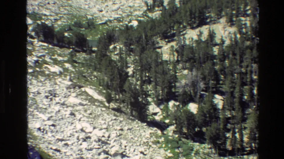 1980 8mm frame from Grand Teton, Wyoming, looking down over pale rock fields, green alpine ground, and clustered evergreens near lake water.