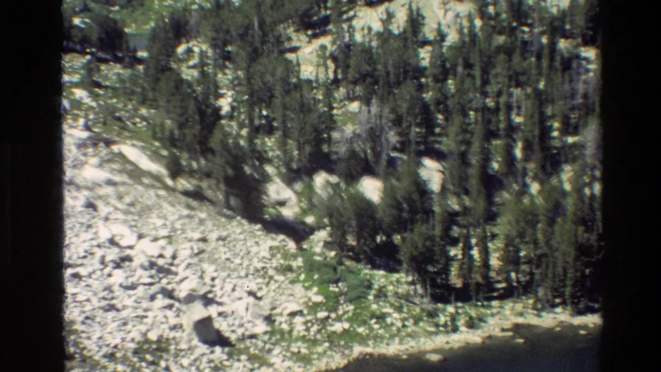 Grand Teton 1980 8mm aerial frame showing conifers, scattered boulders, rocky alpine ground, and a curved lake shoreline below.