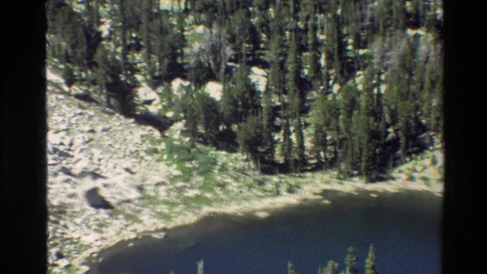 1980 8mm Grand Teton aerial view with dark lake water in the foreground, dense conifers, pale rocks, and green alpine terrain beyond.