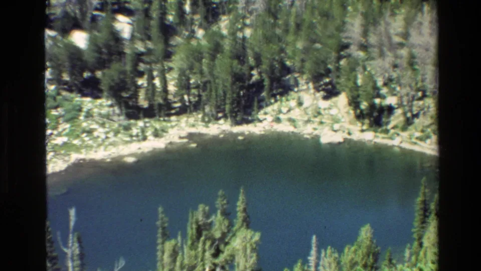 Grand Teton, Wyoming home-movie frame with a curved alpine lake edge, evergreen forest, and rocky bank.