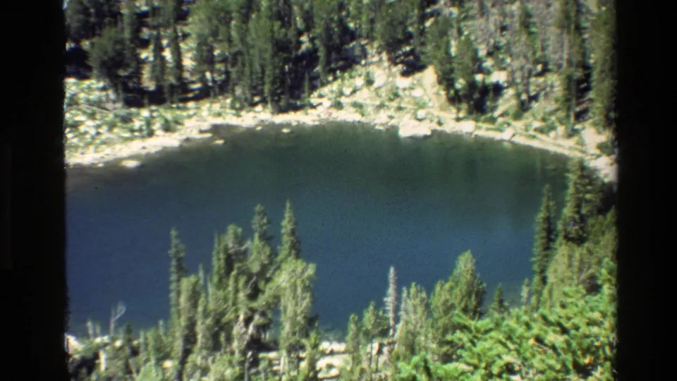 8mm frame from Grand Teton showing dark alpine water, evergreen shoreline, and rocky ground in bright sun.