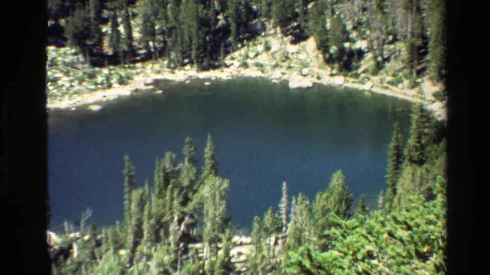 Grand Teton 8mm home-movie view of a dark blue lake ringed by conifers and rocky shoreline.