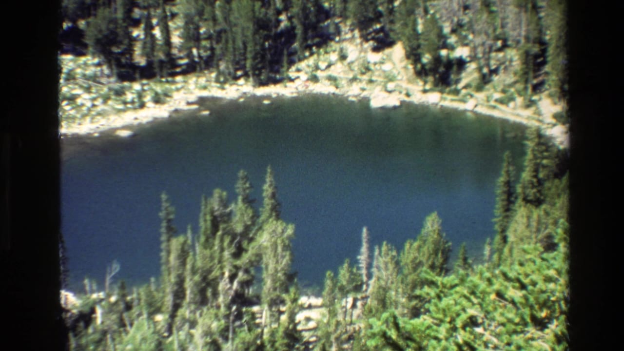 1980 8mm Grand Teton footage showing a blue alpine lake below forested slopes and a pale rocky shore.