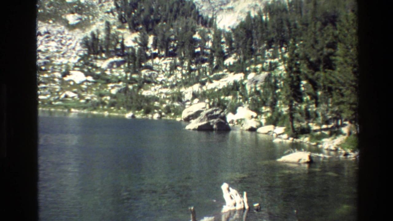 1980 Grand Teton home-movie frame showing a dark lake bordered by conifers, boulders, and steep rocky slopes.