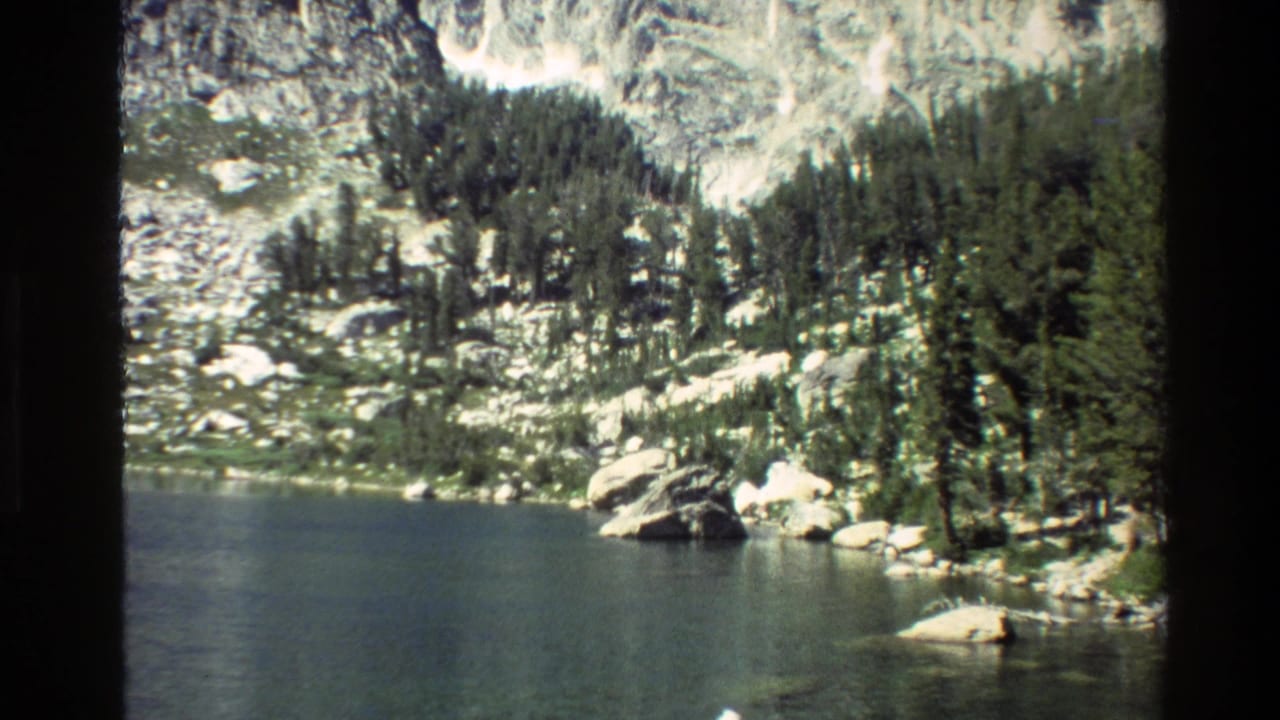 Mountain lake shoreline with dense conifers, boulders, and pale cliffs.