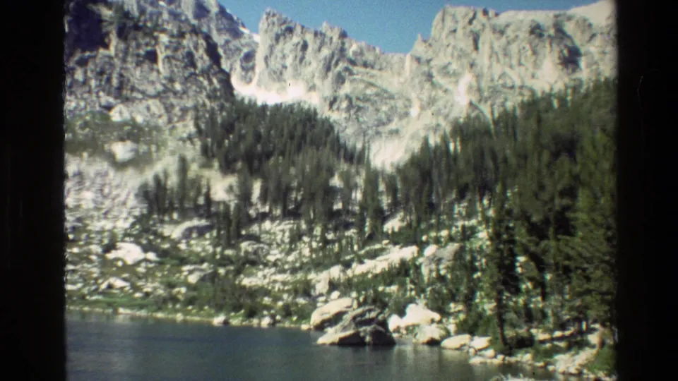 8mm frame from the Grand Teton area showing steep pale rock, dark evergreens, shoreline boulders, and lake water.