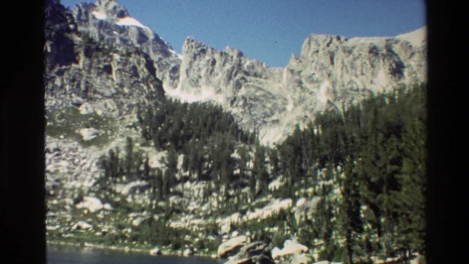Grand Teton 8mm scenic frame with dark lake water, dense conifers, bright stone cliffs, and a clear blue sky.