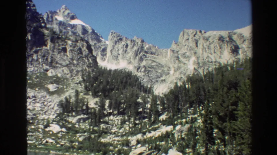 1980 8mm Grand Teton view of rugged snow-streaked peaks rising above a dark green pine forest under blue sky.