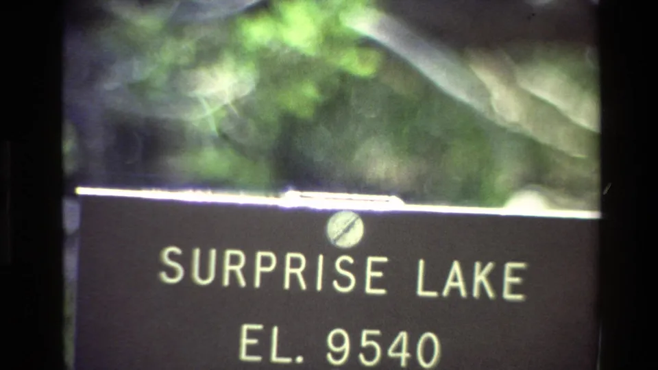 Vintage 8mm close-up of the Surprise Lake sign and elevation text, framed by blurred vegetation and film-edge darkness.