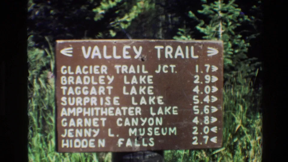 Grand Teton Valley Trail sign with white painted destinations and mileages, surrounded by grass and conifer foliage in 8mm footage.