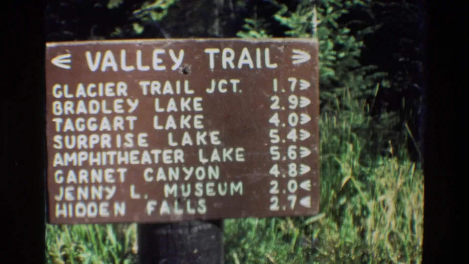 Close 8mm frame of the Valley Trail sign, with readable mileage rows for Bradley Lake, Taggart Lake, Surprise Lake, and other landmarks.