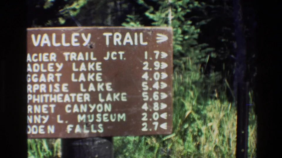 Grand Teton 1980 home-movie frame showing a wooden Valley Trail directional sign with listed distances and green vegetation behind it.