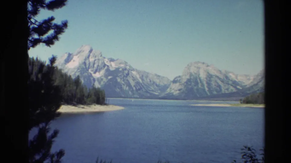A visitor-shot Grand Teton lake view shows blue water, a wooded shoreline, and snow-streaked mountain peaks under a clear sky.