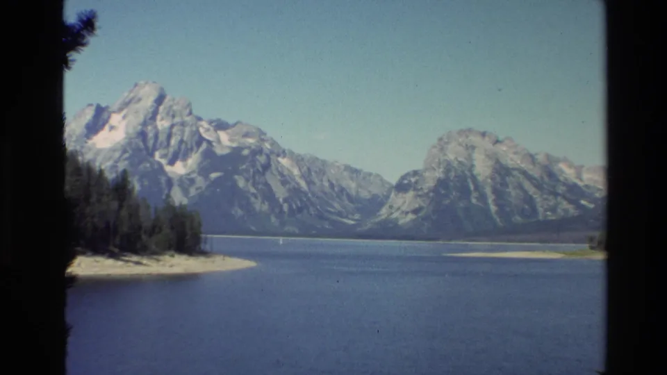 The Grand Teton range appears across a wide blue lake, with evergreen shoreline and pale sky captured in soft 8mm color.
