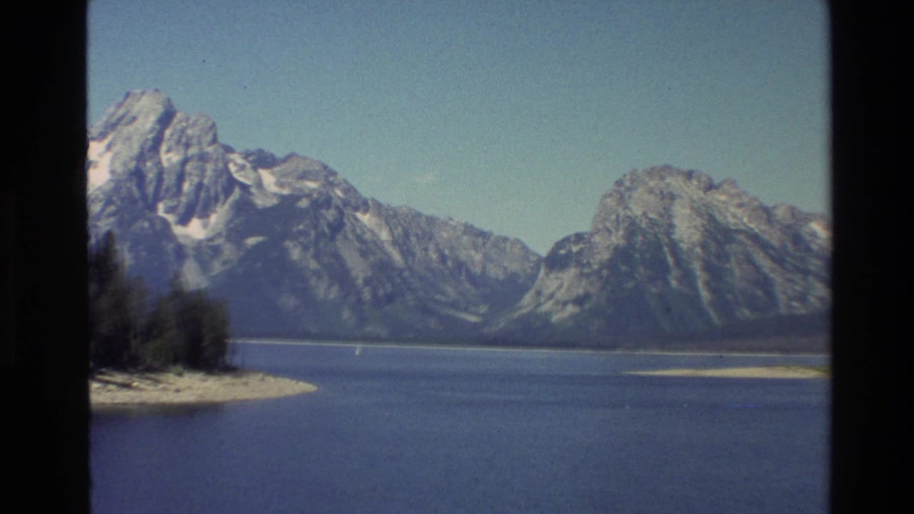 1980 8mm view of the Grand Teton range across a blue lake, with a sandy spit, clear sky, and dark border edges.