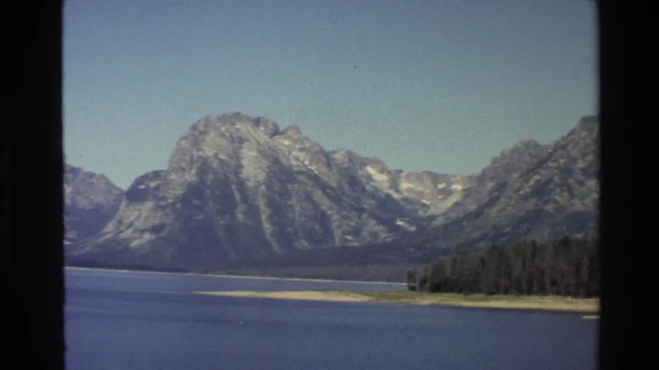 1980 8mm Grand Teton lake view with a rocky peak, evergreen shoreline, pale sandbars, and soft film grain.