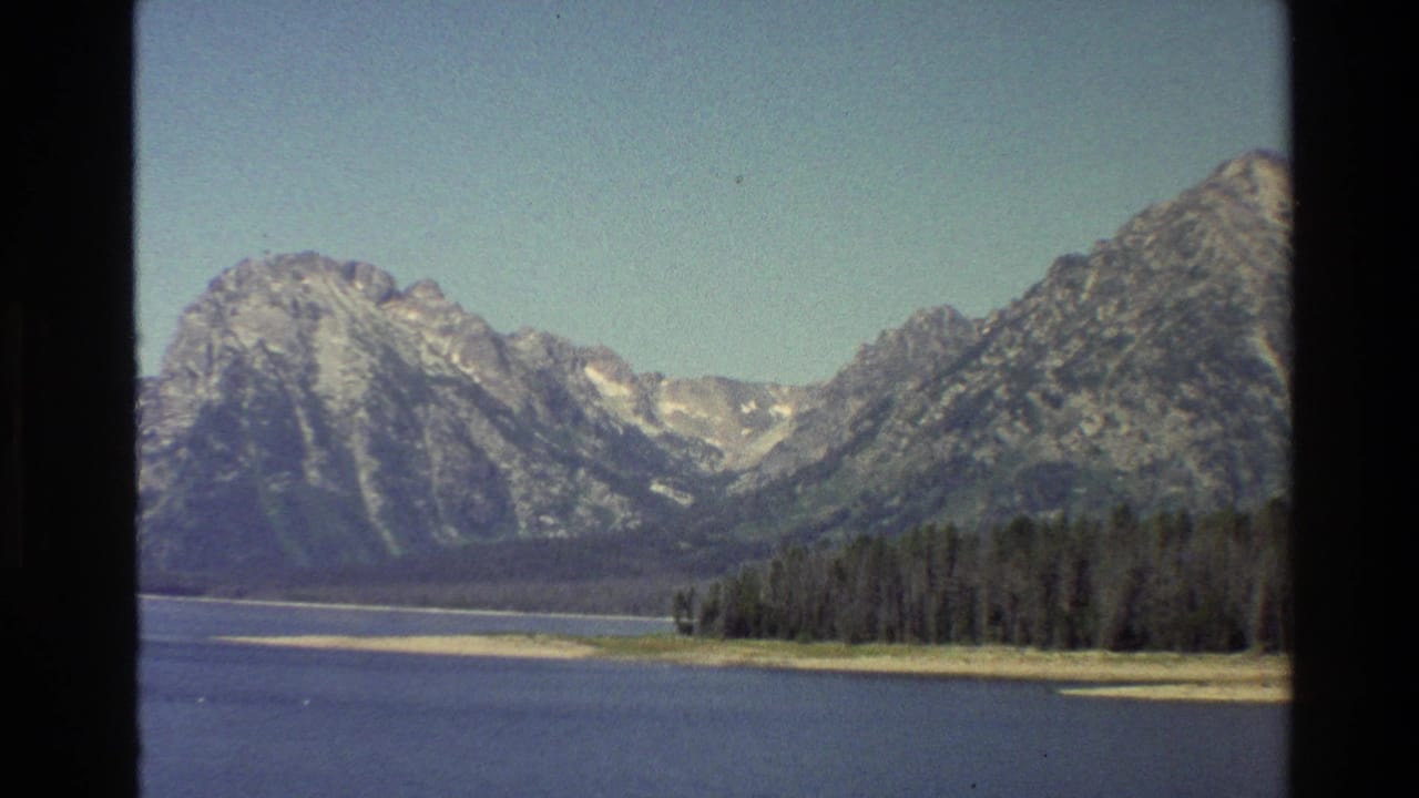 1980 Grand Teton home-movie view of a calm lake, forested shore, and rugged snow-streaked mountains framed by 8mm film edges.