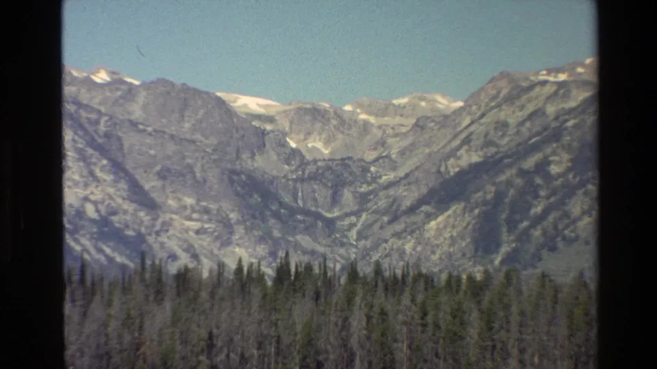 Closer 8mm view of Grand Teton mountains above a dense evergreen shoreline and lake edge.