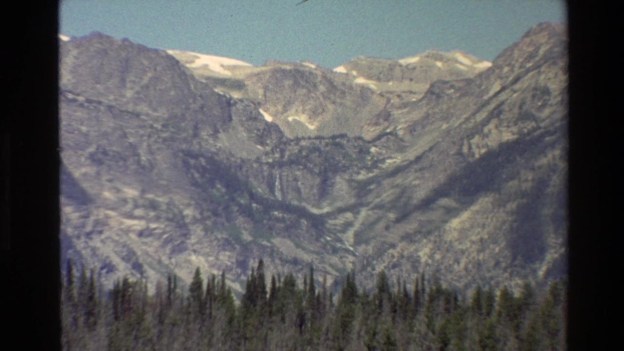 1980 8mm Grand Teton view showing rocky peaks, small snow patches, blue sky, and a dark evergreen forest line.