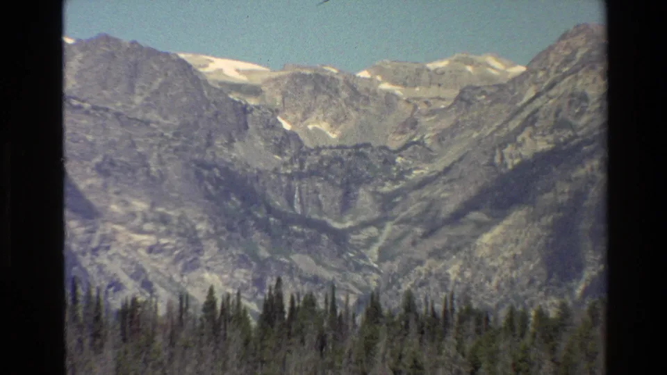 1980 8mm Grand Teton view showing rocky peaks, small snow patches, blue sky, and a dark evergreen forest line.