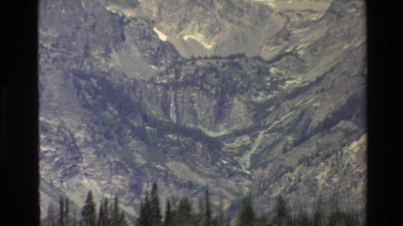 Closer mountain view with steep gray rock faces, snow patches, forest below, and dark frame edges.