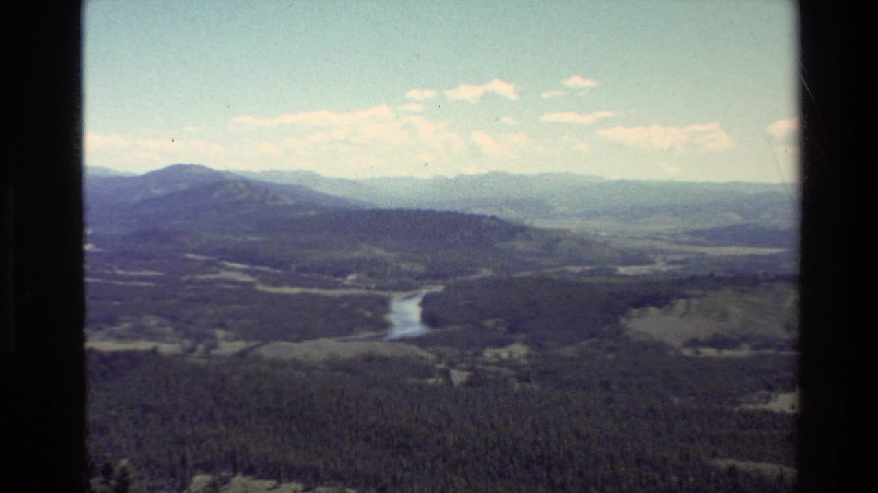 1980 8mm view over a forested Grand Teton-area valley, with a pale river bend, open fields, distant mountain ridges, and scattered clouds.