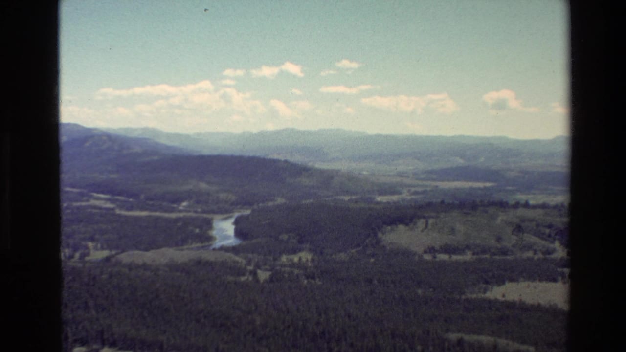 Wide valley landscape with river curve, dark forest, distant mountains, clouds, and film gate edges.