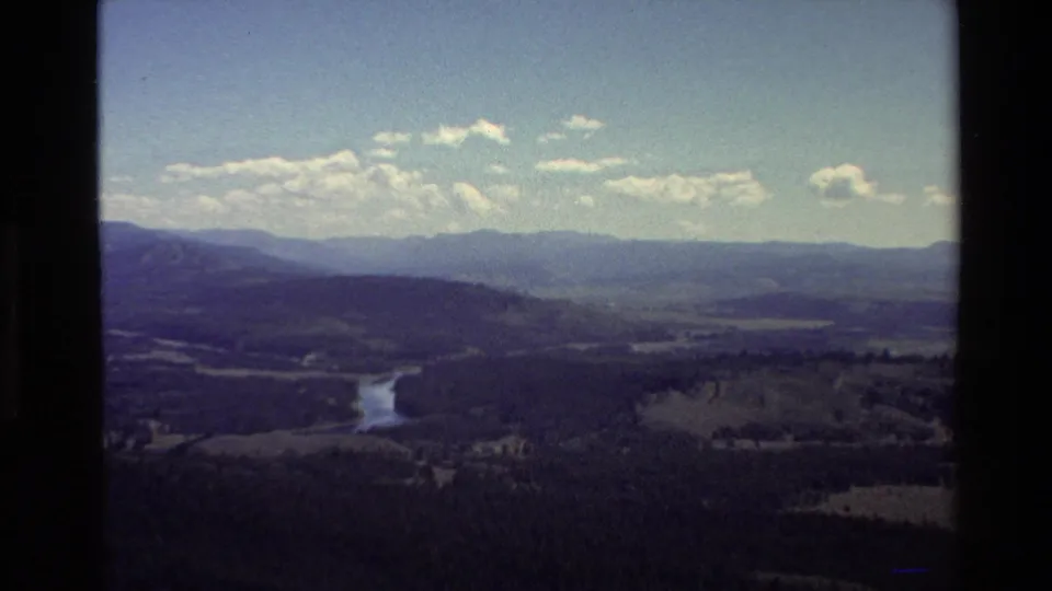 Soft 8mm frame from 1980 showing an elevated view of a Grand Teton-area forest valley, winding river, distant ridgeline, and scattered white clouds.