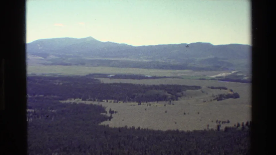 1980 home-movie frame of the Grand Teton, Wyoming landscape with meadow-like valley floor, forest patches, water, and mountains in the distance.