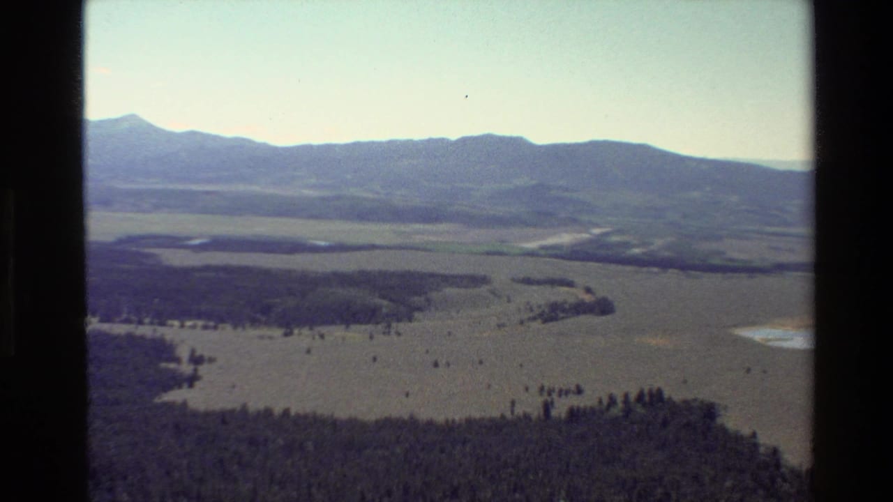 1980 8mm aerial view near Grand Teton, Wyoming, showing open plains, dark forest areas, small water bodies, and distant mountains.