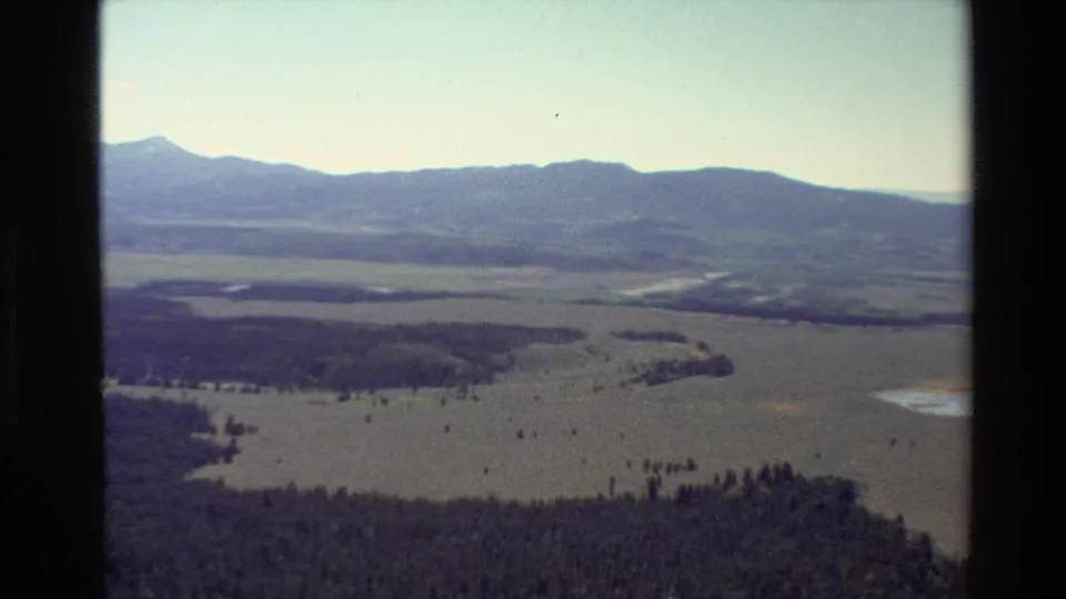 1980 8mm aerial view near Grand Teton, Wyoming, showing open plains, dark forest areas, small water bodies, and distant mountains.