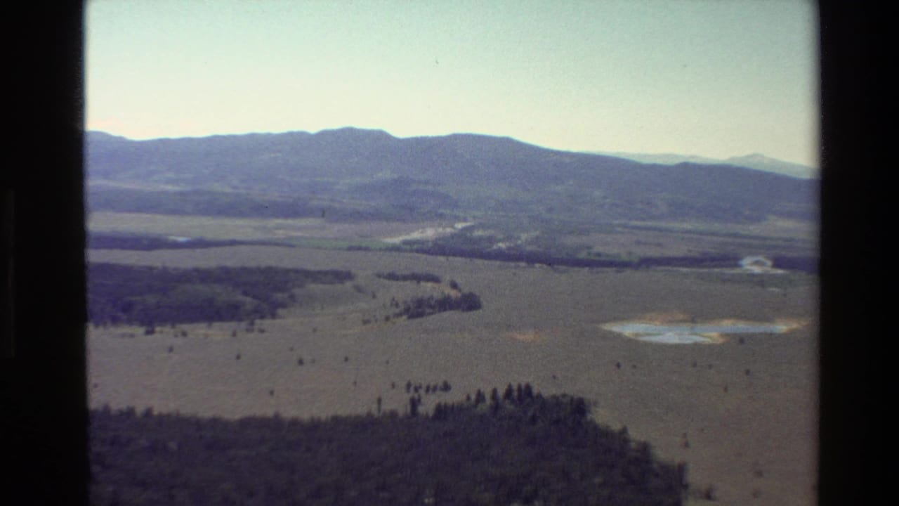 Wide aerial landscape with mountain ridge, plains, forest patches, and reflective water areas.
