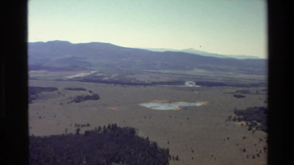 1980 8mm aerial frame over Grand Teton area terrain, with foreground forest, broad plain, water patches, and distant ridgelines.