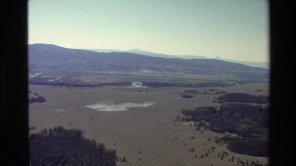 Grand Teton, Wyoming aerial home-movie frame from 1980 showing broad plains, tree clusters, water patches, and distant mountain forms.