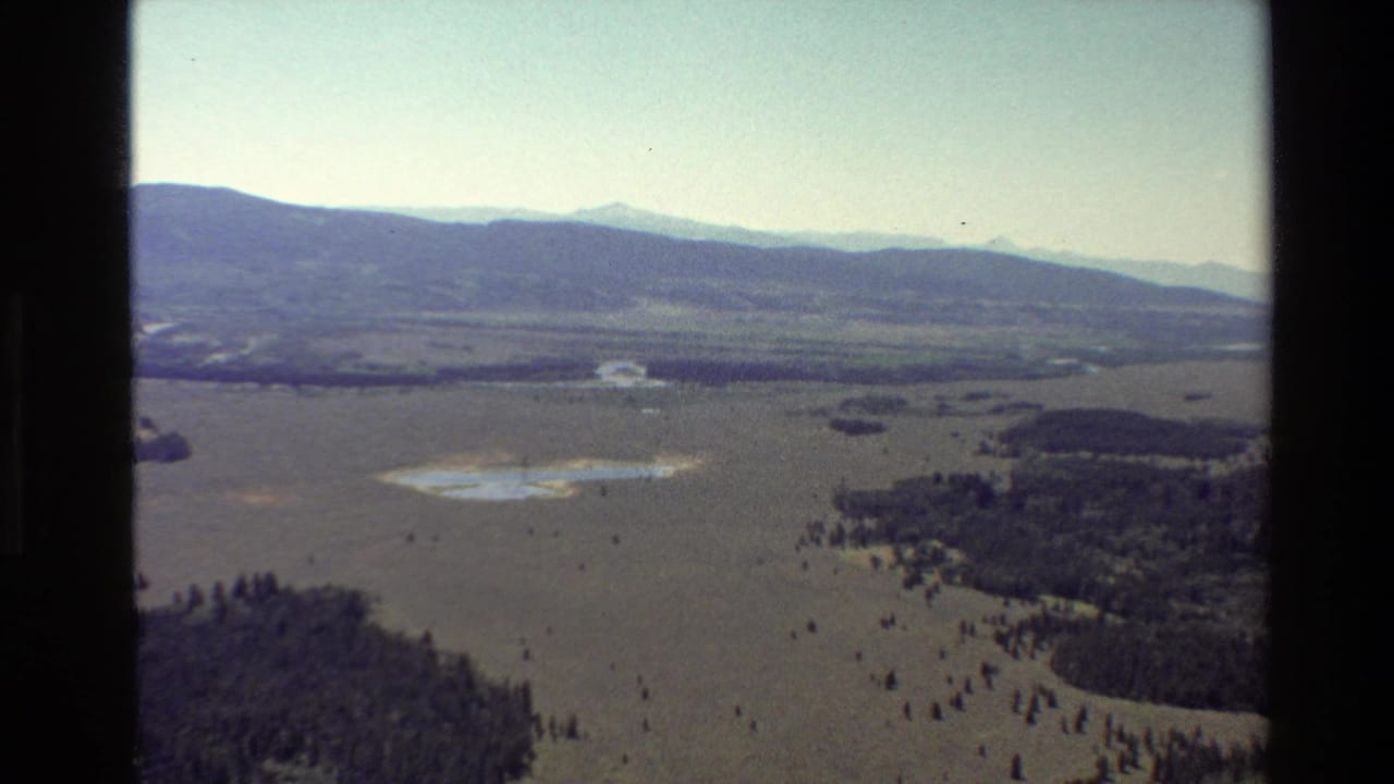 Aerial landscape with mountains, open valley floor, trees, and small reflective water patches.
