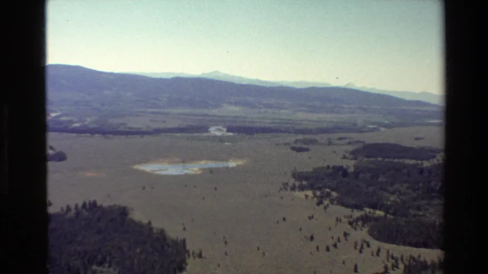 Aerial 8mm frame over the Grand Teton area, with a wide valley floor, dark tree clusters, pale water patches, and layered mountains beyond.