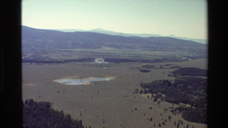 Vintage 8mm aerial view in Grand Teton, Wyoming, showing wetland-like water patches, an open valley, scattered trees, and a soft mountain ridge.