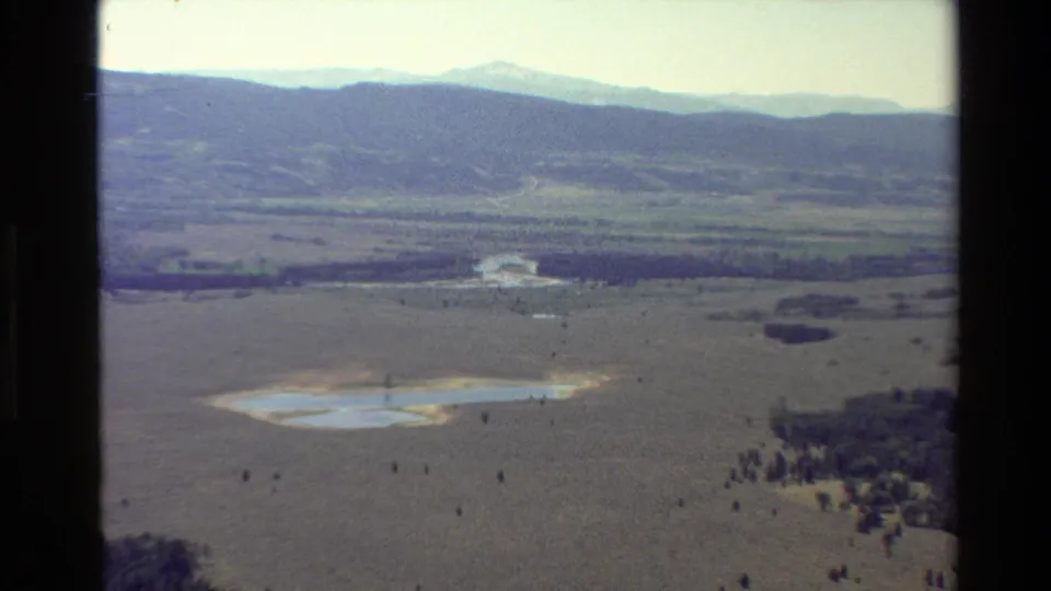 1980 aerial home-movie frame over Grand Teton, Wyoming, with a small lake or wetland, open meadow, wooded edges, and hazy mountain peaks.