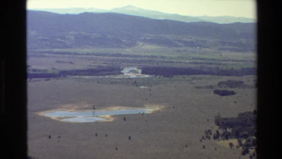 1980 8mm aerial view in the Grand Teton, Wyoming area showing a dry valley, small reflective lake, sparse trees, and hazy mountain ridges.