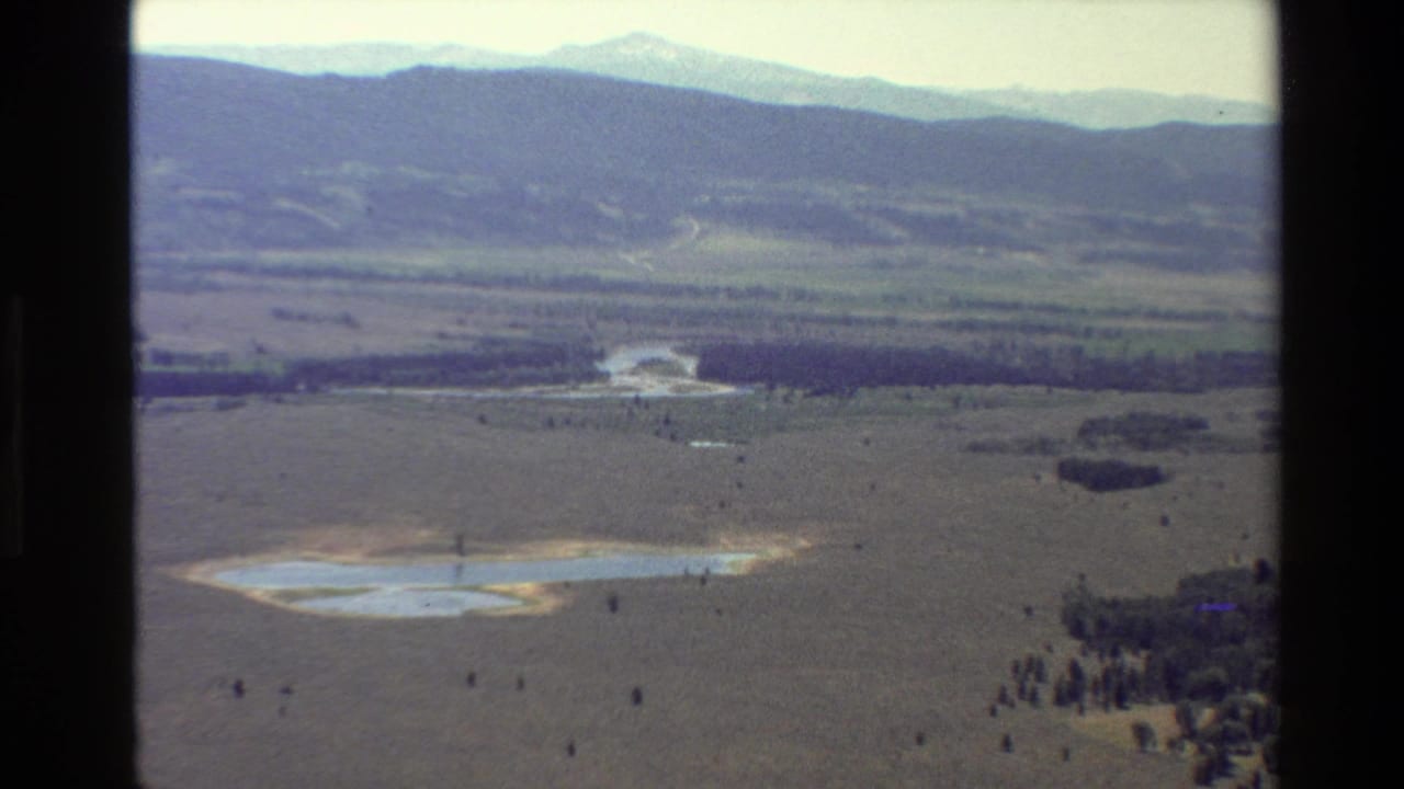 Wide aerial landscape with a reflective lake, open flat terrain, tree clusters, and mountains beyond.