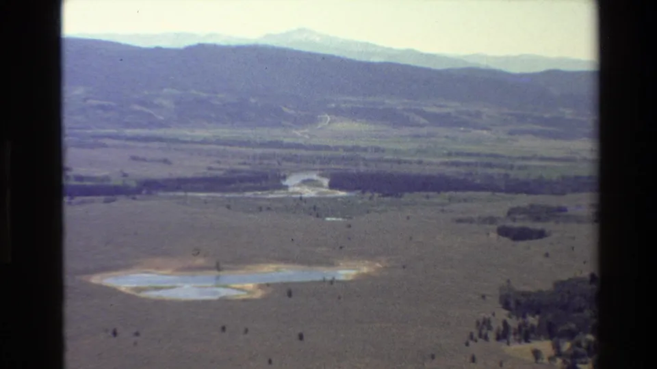 1980 8mm aerial view near Grand Teton with a muted valley floor, small pale lake, dark tree line, and hazy mountains in the distance.