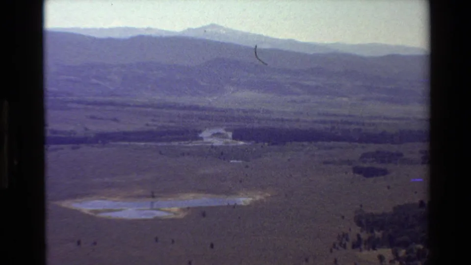 Faded 1980 8mm frame from the Grand Teton area showing a broad valley, pale lake, scattered trees, and distant mountain ridges.