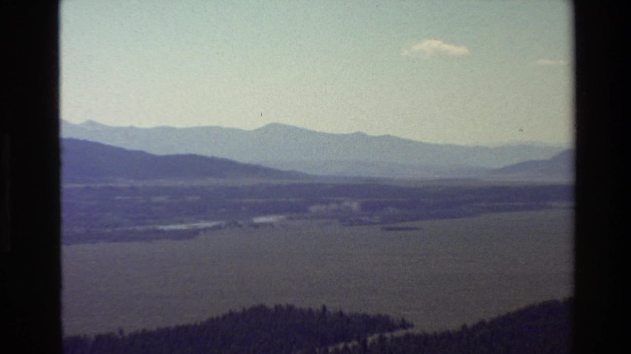 1980 8mm Grand Teton, Wyoming vista with a broad lake, valley floor, forested foreground, and hazy mountain ridges.