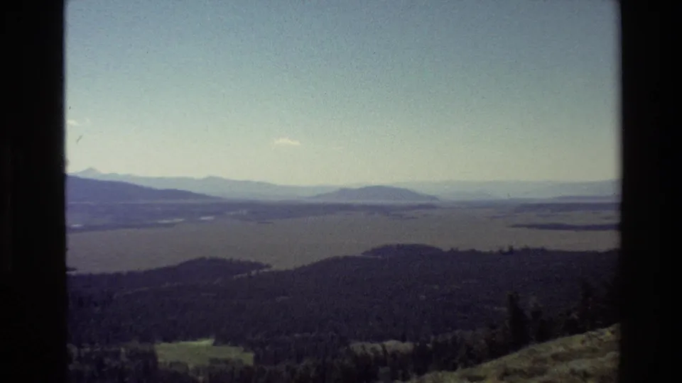 1980 Grand Teton, Wyoming overlook view with lake water, evergreen hills, valley fields, and hazy mountains in the distance.