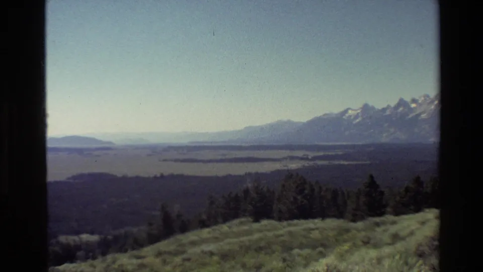 Grand Teton landscape view from 1980 home-movie footage, with lake flats, wooded hills, and snow-dusted peaks across the horizon.